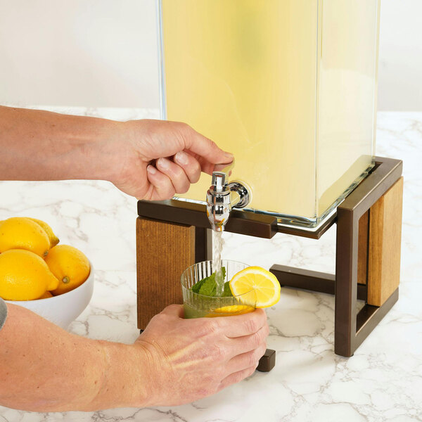 A person pouring water from a Cal-Mil Sierra Bronze and Rustic Pine beverage dispenser into a glass.