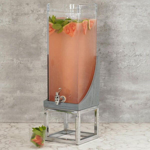A Cal-Mil Ashwood Gray Oak wood beverage dispenser with watermelon slices and mint leaves with a glass of pink liquid next to it.