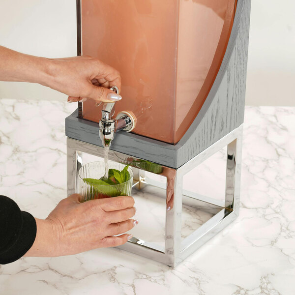 A person filling a glass with a drink from a Cal-Mil Ashwood Gray Oak Wood Beverage Dispenser.