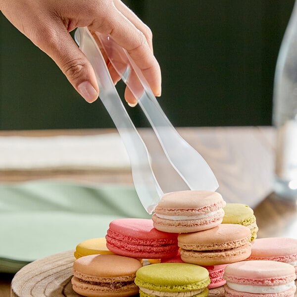 A pair of clear disposable plastic tongs being used to pick up colorful macarons.