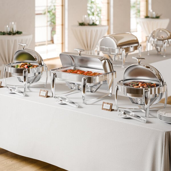 A buffet table with three shiny stainless steel chafing dishes containing food, set on a white tablecloth with serving utensils and small label cards in front.