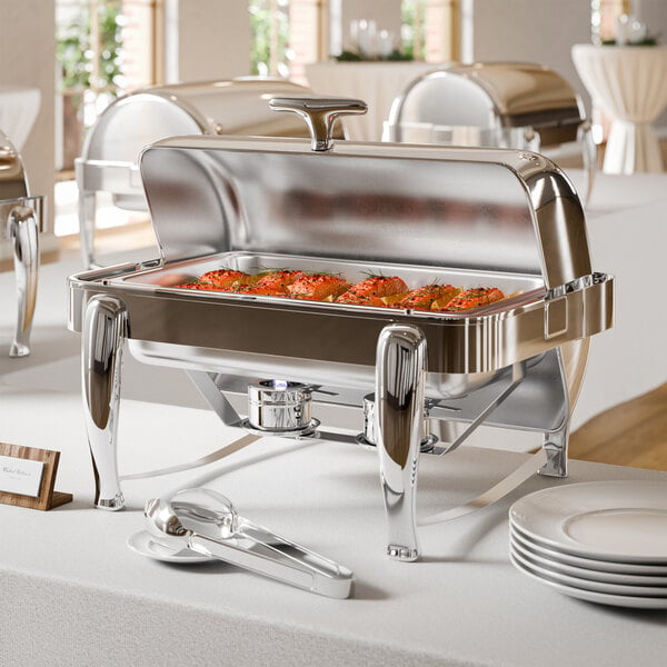 A stainless steel chafing dish with a partially open lid, containing food, is displayed on a white tablecloth with serving tongs and stacked plates nearby.
