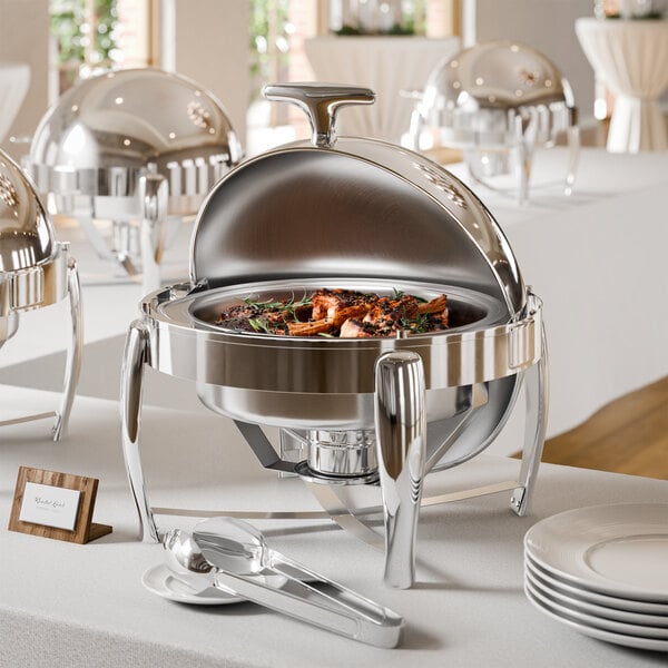 A shiny, round stainless steel chafing dish with a partially open lid, containing cooked food, is displayed on a white tablecloth with serving utensils and plates nearby.