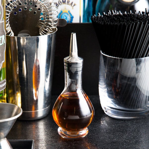 A clear glass American Metalcraft bitters bottle on a table next to a glass of brown liquid.