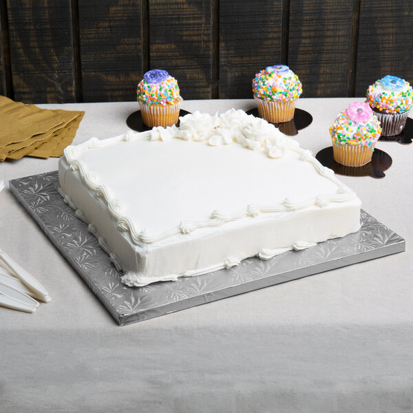 A white cake on a silver square cake drum on a table with cupcakes on top.