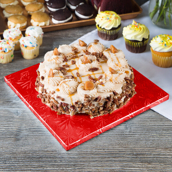A cake on a red Enjay cake drum on a table with cupcakes.