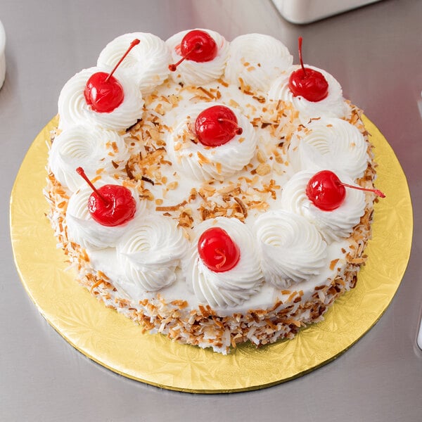 A white frosted cake with red cherries on a gold cake board.