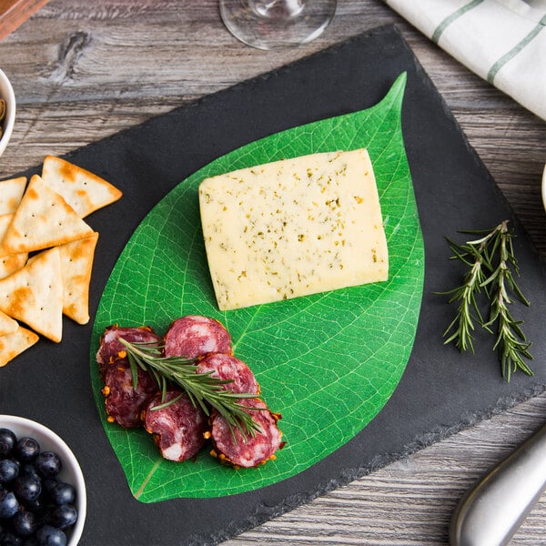 Black charcuterie board with cheese and sliced meat on green leaf, crackers, berries, and herbs