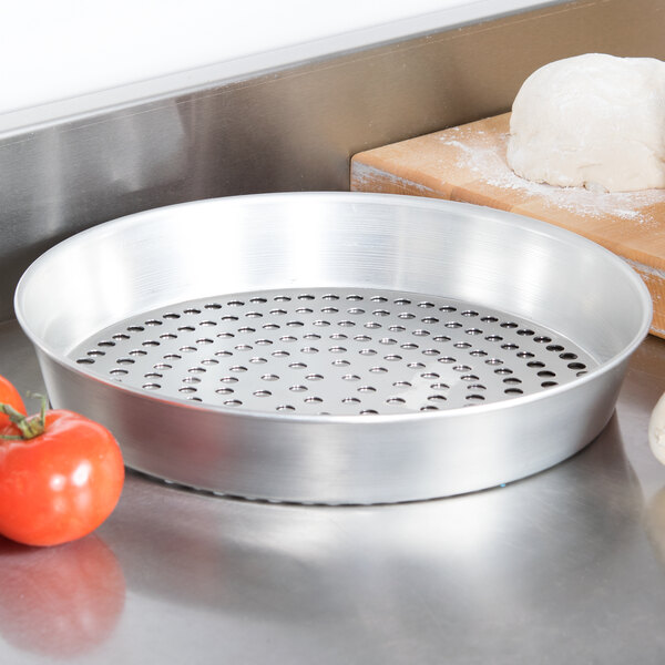 An American Metalcraft silver tin-plated steel pizza pan with holes in it sitting on a table with a ball of dough and tomatoes.