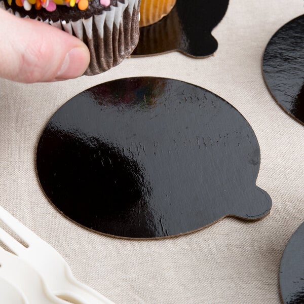 A hand holding a cupcake with a black and brown Enjay round cake board with a tab.
