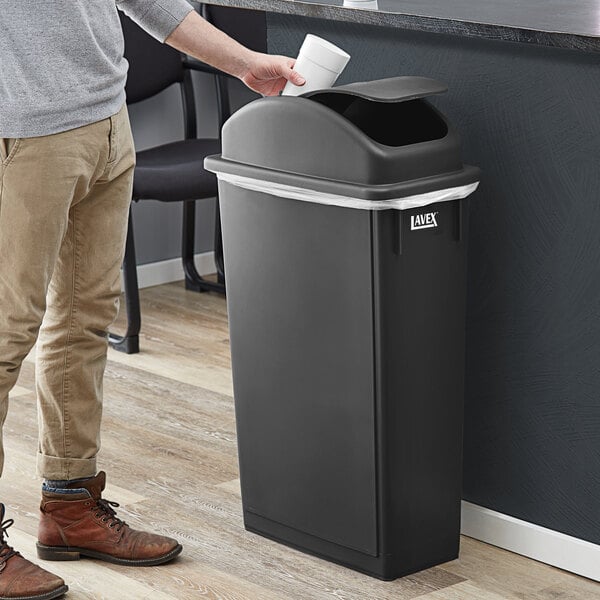 A man throwing a white cup into a black Lavex Slim Rectangular Trash Can with a swing dome lid.