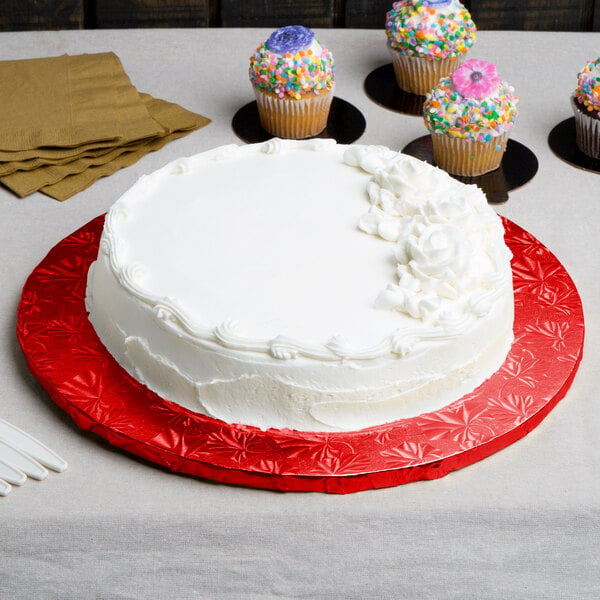 A white frosted cake on a red Enjay round cake drum with cupcakes on a table.