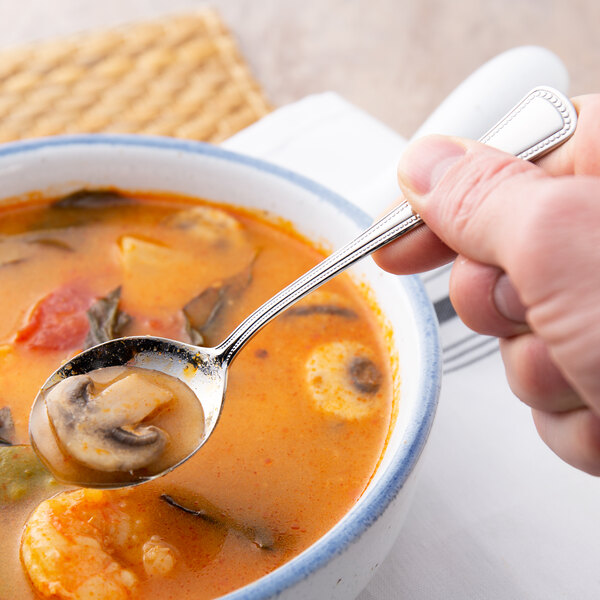 A person holding a Oneida Needlepoint stainless steel bouillon spoon over a bowl of soup.