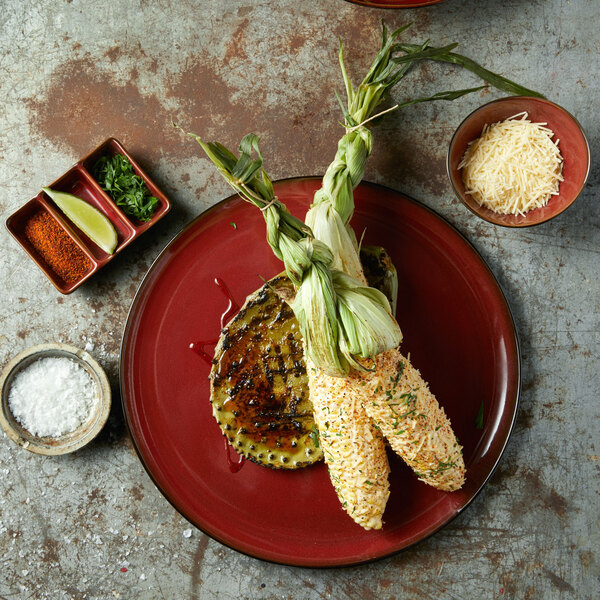 A Oneida Rustic crimson porcelain pizza plate with a lime wedge and spices on a table with a plate of grilled corn.