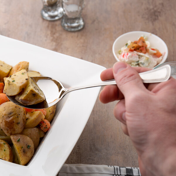 A person holding a Oneida Needlepoint stainless steel serving spoon over a plate of food.