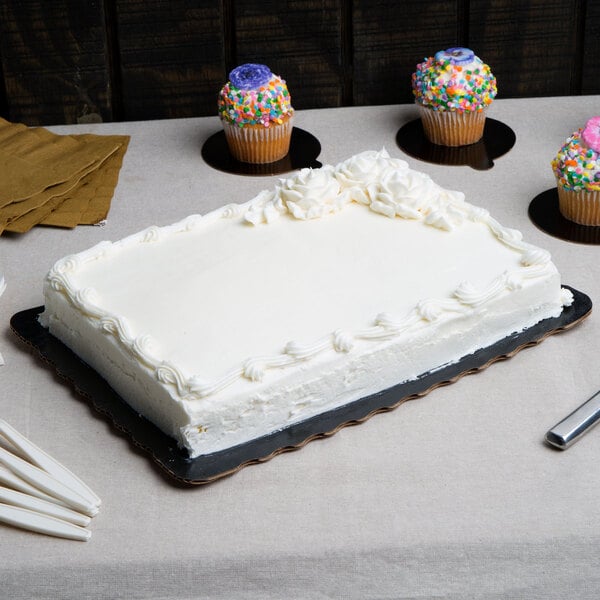 A white cake with frosting on a black Enjay laminated corrugated sheet cake board on a table with cupcakes.