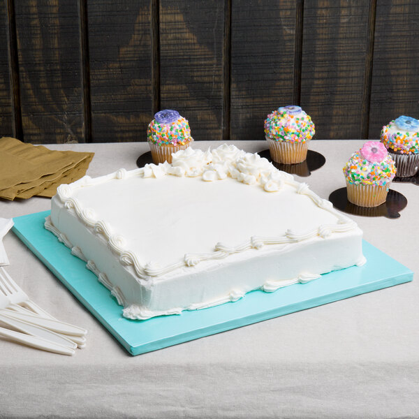 A white cake with frosting on a blue Enjay square cake drum on a table with cupcakes and a donut.