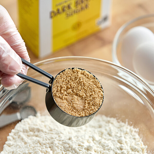 A gloved hand uses a measuring cup to scoop brown Domino Dark Brown Sugar.