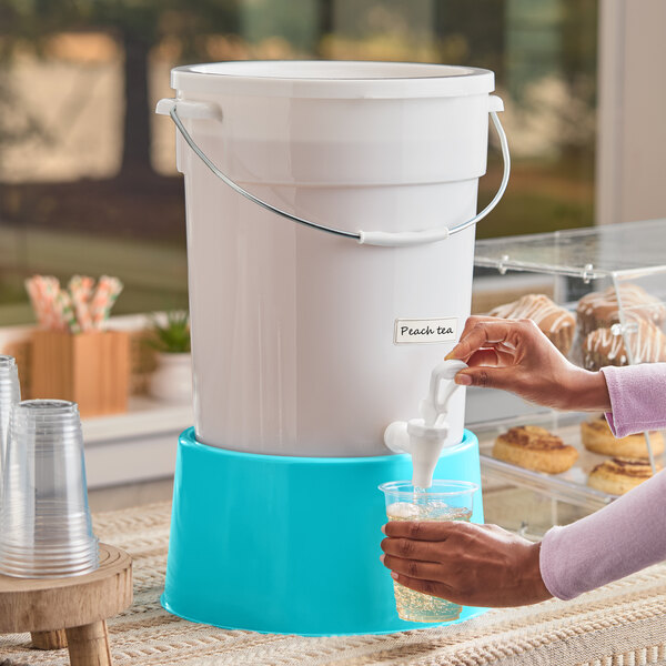 A large white beverage dispenser with a blue base and a spigot, labeled for peach tea, being used to fill a clear plastic cup.