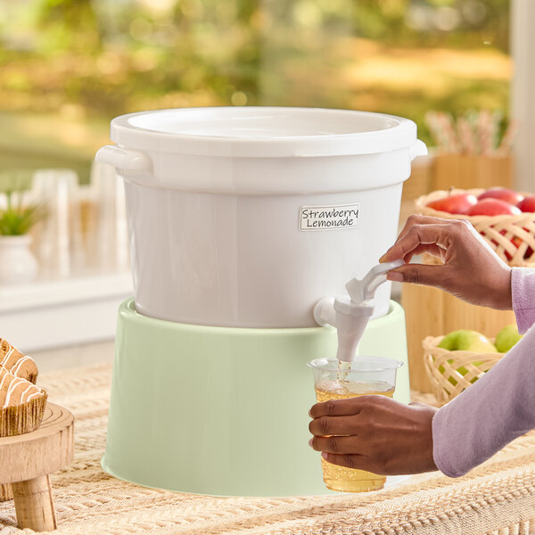 A white round beverage dispenser with a spigot and a light green base, being used to pour a drink into a cup.