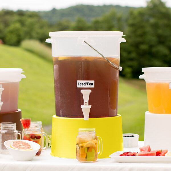 A Choice translucent plastic beverage dispenser with a yellow base on a table with a pitcher of fruit and water.