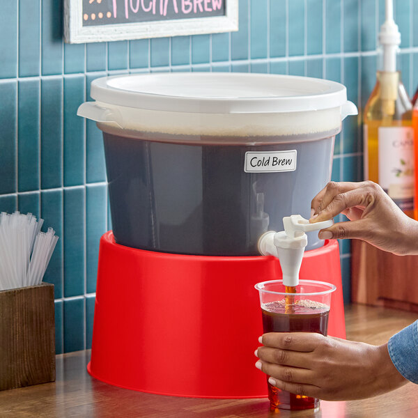 A round 3-gallon translucent beverage dispenser with a red base and a white lid, being used to pour cold brew coffee into a cup.