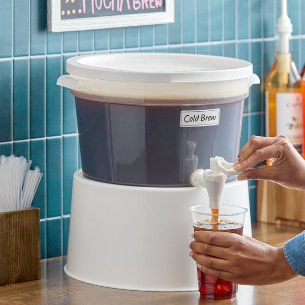 A round, translucent 3-gallon beverage dispenser with a white base and lid, being used to pour cold brew coffee into a cup.
