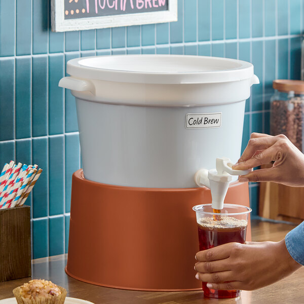 A white round beverage dispenser with a brown base being used to pour cold brew into a plastic cup.