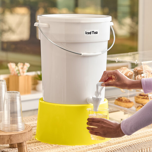 A large white beverage dispenser labeled 'Iced Tea' with a yellow base, being used to pour a drink into a clear cup.