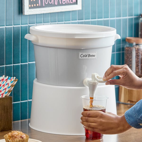 A white 3-gallon round beverage dispenser with a white base being used to pour cold brew into a plastic cup.