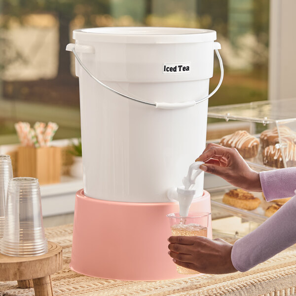A large white beverage dispenser with a rose-colored base labeled 'Iced Tea,' being used to pour a drink into a cup.