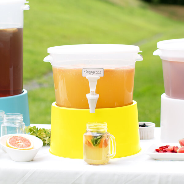 A Choice translucent plastic beverage dispenser with yellow base on a table with cups and a glass jar of pink liquid.