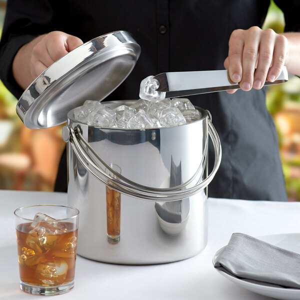 A woman using tongs to put a silver lid on a Tablecraft stainless steel ice bucket.