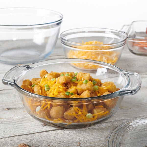 A clear glass round cake pan with handles, filled with food and accompanied by other empty glass bowls.