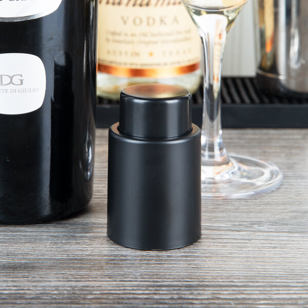 A black Franmara wine pump and stopper on a wood surface next to a bottle of wine.