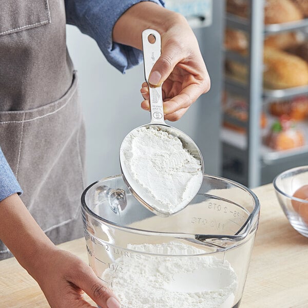 A person using a Vollrath stainless steel measuring cup to scoop white powder.