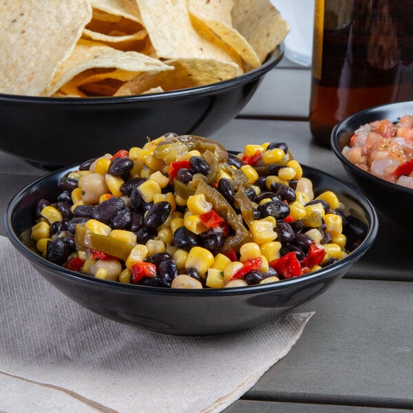 A black Vollrath melamine bowl filled with black bean and corn salsa on a table with chips and a bottle of beer.