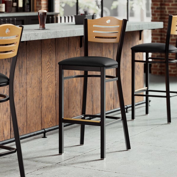 A Lancaster Table & Seating black wood bar stool with a black vinyl seat and natural wood back.