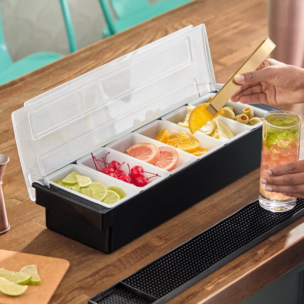 A six-compartment condiment bar filled with various garnishes such as lime slices, cherries, grapefruit, orange slices, and olives.