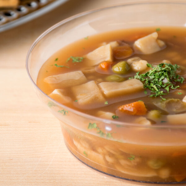 A close-up of a clear Fineline plastic bowl filled with soup, vegetables, and meat.