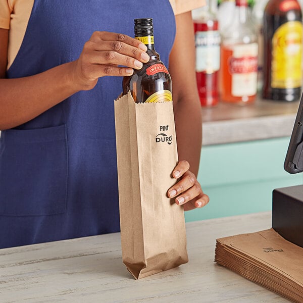 A woman's hand holding a Duro brown paper bag with a bottle of alcohol inside.