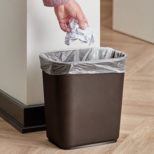 A hand throws a crumpled paper into a brown Continental rectangular trash can.