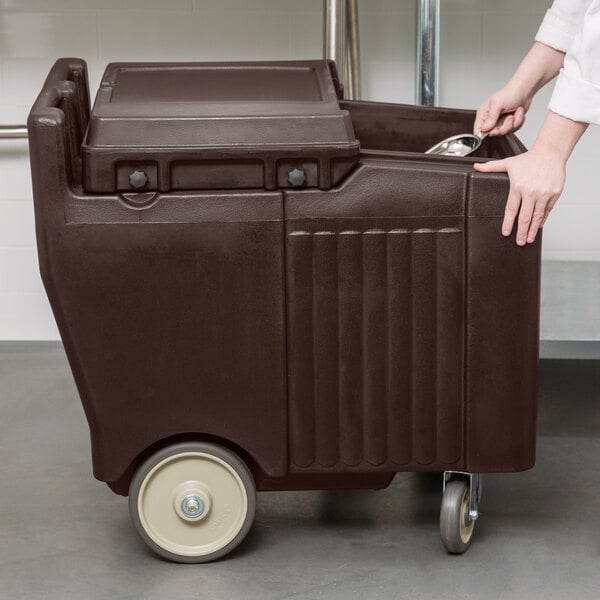 A woman pushes a dark brown Cambro mobile ice bin on wheels.