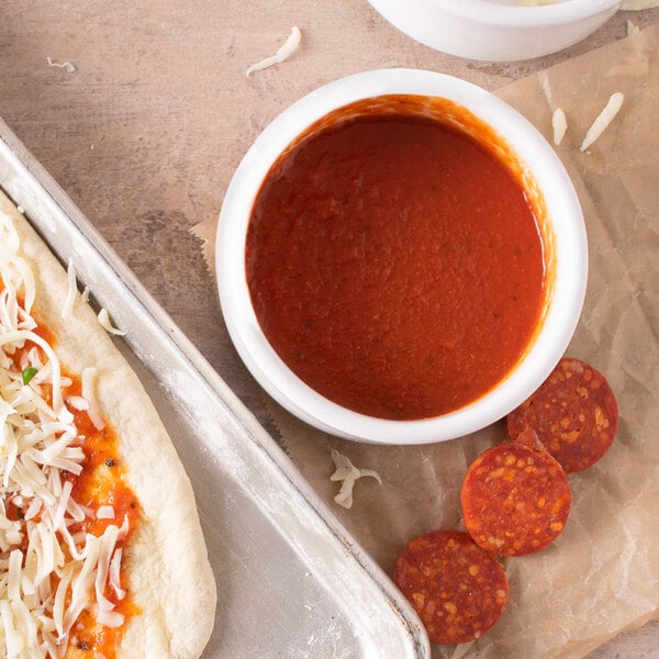 small bowl of marinara sauce beside a pizza and pepperoni slices