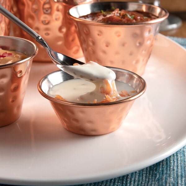 A close-up of a Choice hammered copper stainless steel sauce cup with a spoon pouring white liquid over a bowl of food.