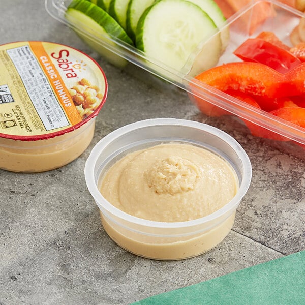 A close up of a plastic container of Sabra Classic Hummus on a table with other food containers.