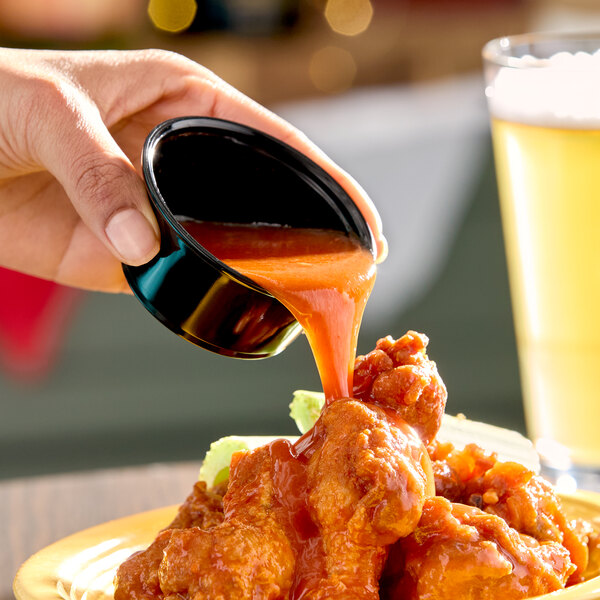 A hand is pouring Frank's RedHot XTRA Hot Cayenne Sauce from a small black cup onto a plate of chicken wings.
