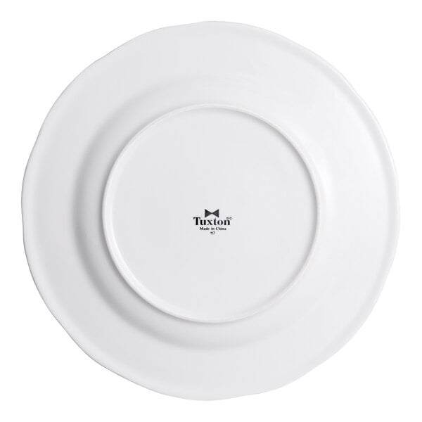 A bright white china plate with a slightly wavy rim, shown from the bottom with the Tuxton brand mark.