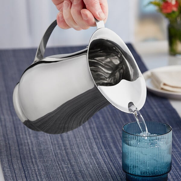 A close-up of a hand pouring water from a silver Acopa pitcher into a glass.