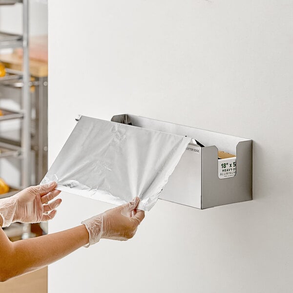A stainless steel film and foil dispenser and cutter mounted on a wall, with a person pulling out a sheet of aluminum foil.